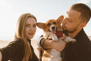 joven pareja feliz con perro toma selfie en la playa. hermosa chica y chico y cachorro corgi foto