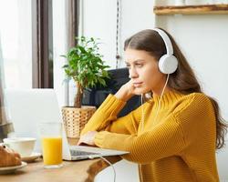 Happy girl in wireless headphones studying online, using laptop and taking breakfast photo