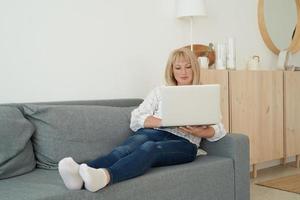 Mature woman using laptop while sitting on comfortable sofa, home interior photo