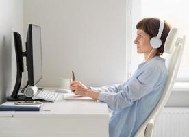 Side view of middle aged woman working remotely from home. Happy female in wireless headphones photo