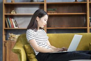 Beautiful Japanese female freelancer smiling and typing at computer in hotel on couch photo