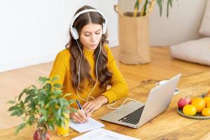Happy female in wireless headphones studying online course, using pc and writing in notepad photo