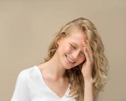 Beautiful young girl laughing. Pretty blonde with curly hair in white t shirt against beige wall photo