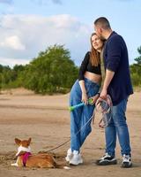 Young happy couple and dog standing on beach against pines and sand, man gently kissing woman photo