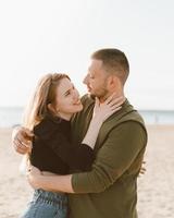 Young adult couple standing on beach, looking each other. Man embracing laughing woman photo