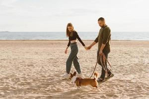 Young happy couple and dog walking along beach. Man holding hand of woman and Corgi puppy