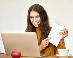 mujer feliz con auriculares hablando por conferencia telefónica y chat de video en una laptop en la oficina
