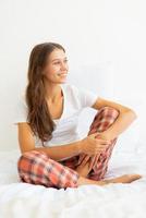 Beautiful young woman without makeup sitting on bed and looking away after waking up. photo