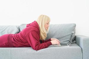 Mature woman working at home with laptop laying on comfortable sofa, room interior. photo