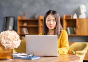 Asian woman working on laptop at home or in cafe. Young lady in bright yellow jumper photo