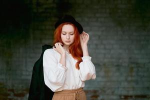 Redhead woman is resting against dark grunge wall. Female in black hat photo