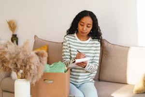 mujer afroamericana que lleva la cuenta de la ropa en una caja de cartón. mudarse y empacar