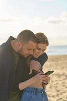 Young couple standing on sandy beach and looking at smartphone, man points finger at mobile photo