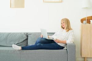 Full length mature woman using laptop laying on comfortable sofa, home interior photo