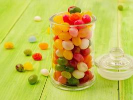 glass jar with lid filled with colorful candies on a wooden green background, many jelly bean scattered on the table photo