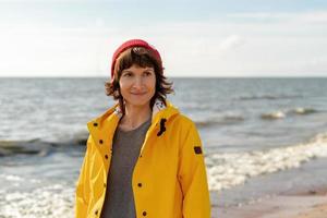 Waist portrait of mature woman in bright yellow cloak and red hat on shore photo