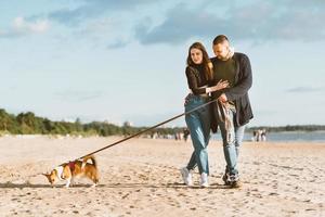 joven pareja feliz y perro caminando en la playa. hombre guapo abrazando suavemente a una mujer hermosa foto