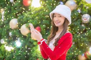 hermosa mujer asiática usa abrigo rojo y sombrero blanco mientras juega al teléfono inteligente en el tema de celebración de navidad y año nuevo. foto
