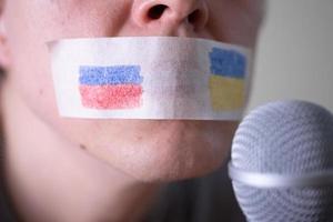 una boca con cinta adhesiva con una bandera de rusia y ucrania tratando de hablar por un micrófono. foto