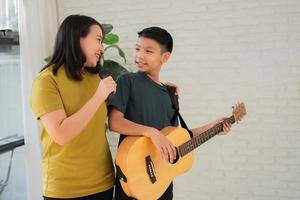 Asian boy playing guitar and mother sing a song and embrace, feel appreciated and encouraged. Concept of a happy family, learning and fun lifestyle, love family ties photo