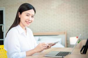 Happy beautiful asian woman working on a laptop and smartphone at the home office sitting at table. Happy female professional freelancer online using notebook pc and smartphone concept. photo