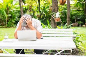 Portrait of an old elderly Asian man holds the head with his hand cause of stress after try to use a computer laptop in the backyard after retired. Concept of Ageism and Hobbies after retirement. photo