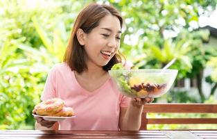 The happy beautiful Asian healthy middle aged woman sitting on the balcony beside the garden and choose between a donut and a vegetable salad. Concept of health care and nutritious food photo