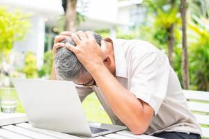Portrait of an old elderly Asian man holds the head with his hand cause of stress after try to use a computer laptop in the backyard after retired. Concept of Ageism and Hobbies after retirement. photo