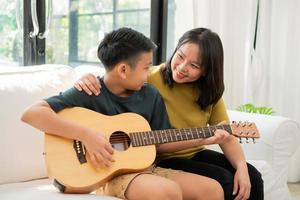 Asian mother embraces son, Asian boy playing guitar and mother embrace on the sofa and feel appreciated and encouraged. Concept of a happy family, learning and fun lifestyle, love family ties