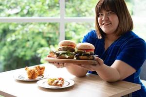 Hungry overweight woman holding hamburger on a wooden plate, Fried Chicken and Pizza on table, During work from home, gain weight problem. Concept of binge eating disorder BED photo