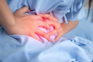 Unhappy Asian patient woman sitting on the hospital bed and holding on stomach suffering. Abdominal pain that comes from menstruation, diarrhea, or indigestion. Sickness and healthcare concept photo