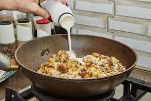 Chef pours cream into pan with fried potatoes, chestnuts, parsley, parmesan on a gas stove. Step by step recipe photo