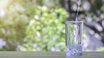 The pure water from jug into glass on wooden table on nature background.