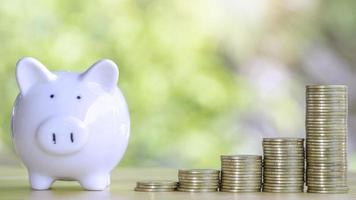 Coins stack 4 rows of gold coins and a white piggy bank placed on a wooden table to save money invest for future. Concept for loan, property ladder, financial, real estate investment, taxes and bonus. photo