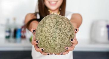 Asian Thai teenager woman holding Cantaloupe Melon on his hand showing and raising up to you with blur modern kitchen background. photo