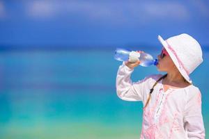 Little girl drinking mineral bottle of water at hot summer day on beach photo