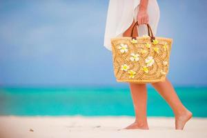 Closeup beautiful bag with frangipani flowers and sunglasses on white beach in female hands photo