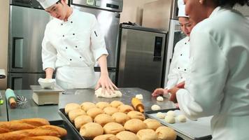 equipe gourmet profissional, três jovens chefs em uniformes e aventais de cozinheiro branco amassam massa de pastel e ovos, preparam pão e alimentos frescos de padaria, assando no forno na cozinha do restaurante de aço inoxidável. video