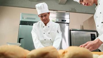 dois chefs asiáticos profissionais em uniformes e aventais de cozinheiro branco estão amassando massa e ovos, preparando pão e alimentos frescos de padaria, assando no forno na cozinha de aço inoxidável do restaurante. video