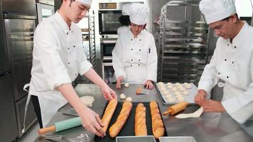 Multiracial professional gourmet team, four chefs in white cook uniforms and aprons knead pastry dough and eggs, prepare bread, and bakery food, baking in oven at stainless steel restaurant kitchen.