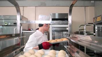 Young professional Asian male chef in white cook uniform with hat, gloves, and apron making bread from pastry dough, preparing fresh bakery food, baking in oven at restaurant stainless steel kitchen. video