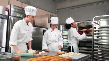 Professional gourmet team, three young chefs in white cook uniforms and aprons knead pastry dough and eggs, prepare bread, and fresh bakery food, baking in oven at stainless steel restaurant kitchen.