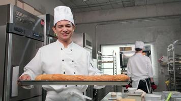Senior Asian male chef in white cook uniform and hat showing tray of fresh tasty bread with a smile, looking at camera, happy with his baked food products, professional job at stainless steel kitchen. video