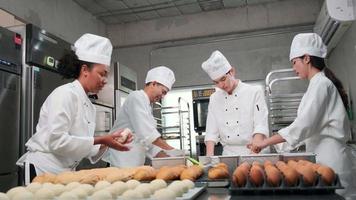 equipo gourmet profesional multirracial, cuatro chefs con uniformes de cocina blancos y delantales amasan masa de pastelería y huevos, preparan pan y comida de panadería, hornean en el horno en la cocina del restaurante de acero inoxidable. video