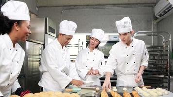 multiraciaal professioneel gastronomisch team, vier chef-koks in witte kookuniformen en schorten kneden deeg en eieren, bereiden brood en bakkerijvoedsel, bakken in de oven in de roestvrijstalen restaurantkeuken. video