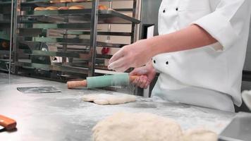 câmera lenta e close-up, chef em uniformes de cozinheiro branco com aventais polvilhe farinha, amassar massa de pastelaria com rolo, preparar pão, tortas e comida de padaria fresca na cozinha de aço inoxidável do restaurante. video