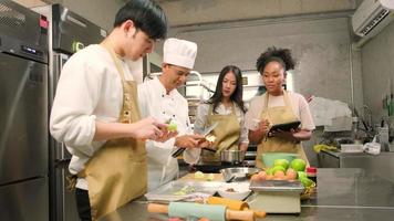 Hobby cuisine course, senior male chef in cook uniform teaches young cooking class students to peel and chop apples, ingredients for pastry foods, fruit pies in restaurant stainless steel kitchen. video