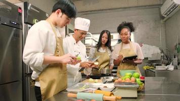 Cours de cuisine passe-temps, le chef masculin senior en uniforme de cuisinier enseigne aux jeunes étudiants en cours de cuisine à éplucher et hacher les pommes, les ingrédients pour les pâtisseries, les tartes aux fruits dans la cuisine en acier inoxydable du restaurant. video