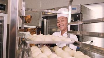 Professional African American female chef in white cook uniform, gloves, and apron making bread from pastry dough, preparing fresh bakery food, baking in oven at restaurant's stainless steel kitchen. video