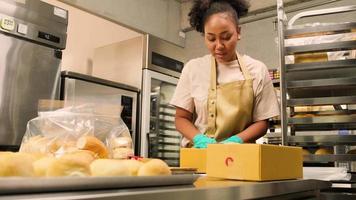 entrega de pequeñas empresas de inicio de panadería. una cocinera afroamericana está empacando pan y pasteles hechos a mano y recién horneados en cajas y enviando para compras de clientes en línea en la cocina culinaria. video